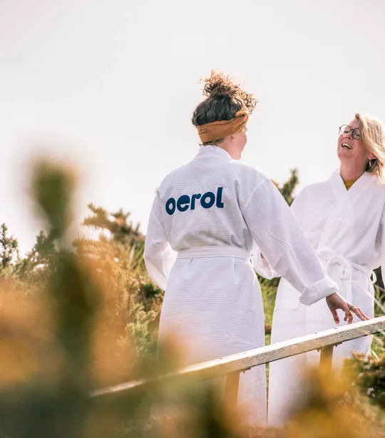 Twee vrouwen dragen een witte badjas met Oerol logo uit de Oerol merchandise collectie van 2023