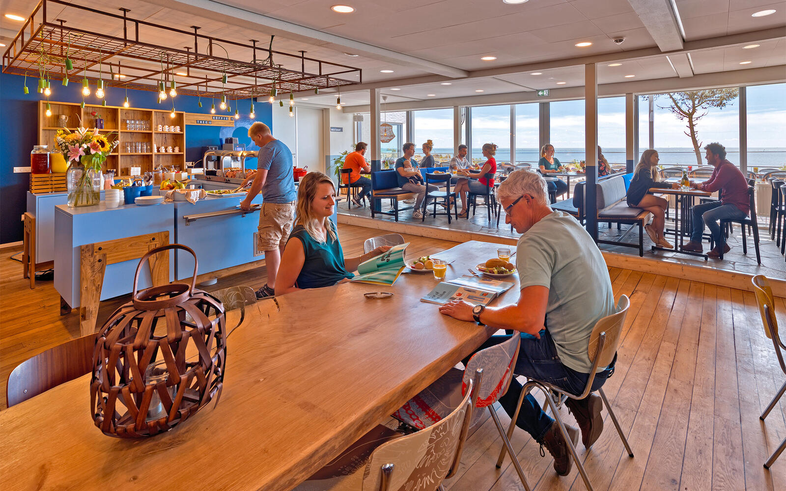 Het met mensen gevulde restaurant van Stayokay Terschelling tijdens het ontbijt, waarbij je door de ramen het uitzicht over het Wad ziet.