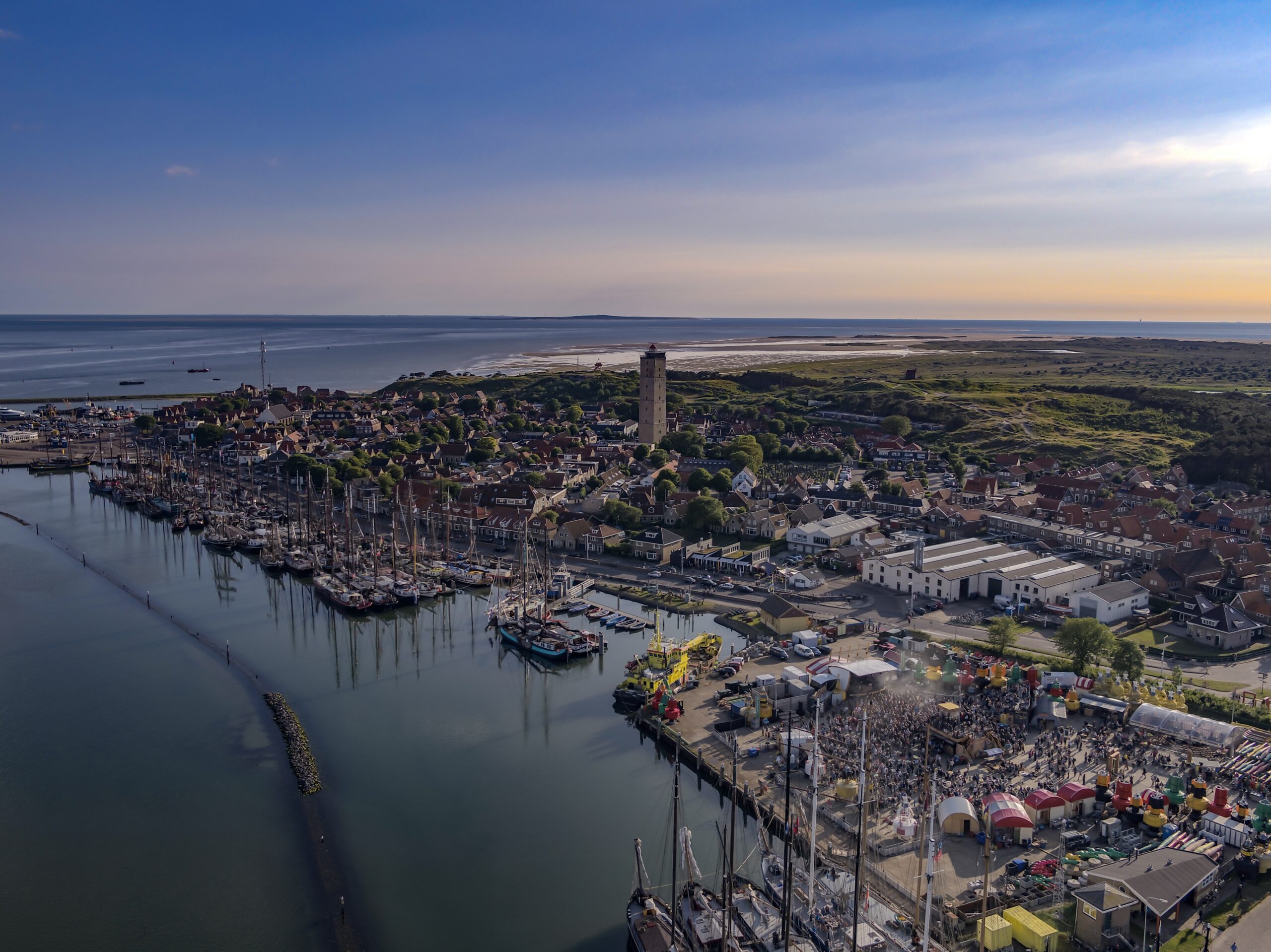 Drone shot van West-Terschelling, waarop je de bruine vloot haven, het dorp, vuurtoren de Brandaris en een met mensen gevuld festivalterrein de Betonning ziet.