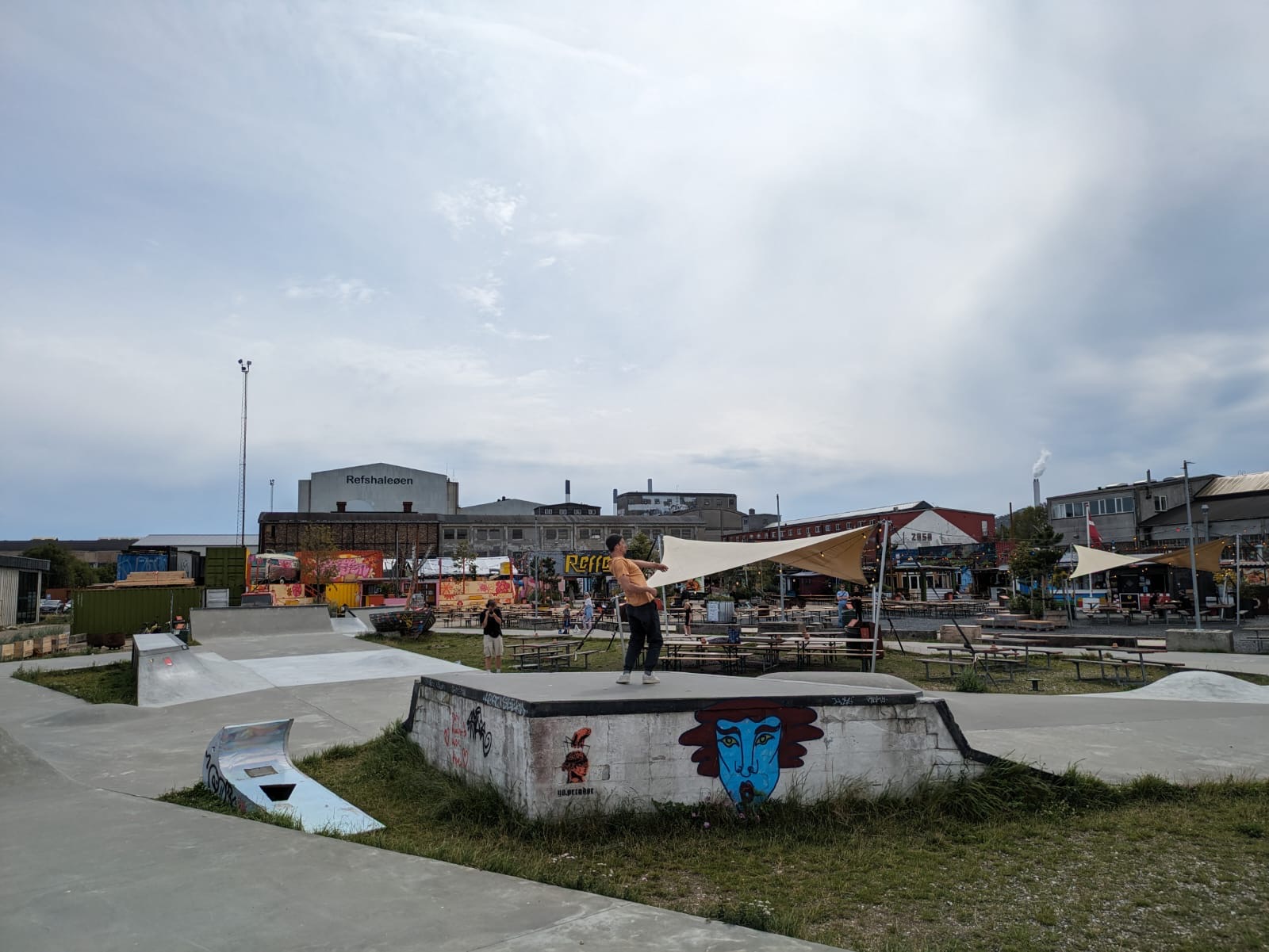 Een overzicht van een kleurrijk, industrieel plein met een skatepark en terrassen. In het midden staat een performer op een verhoging met graffiti, terwijl op de achtergrond mensen rondlopen of zitten.