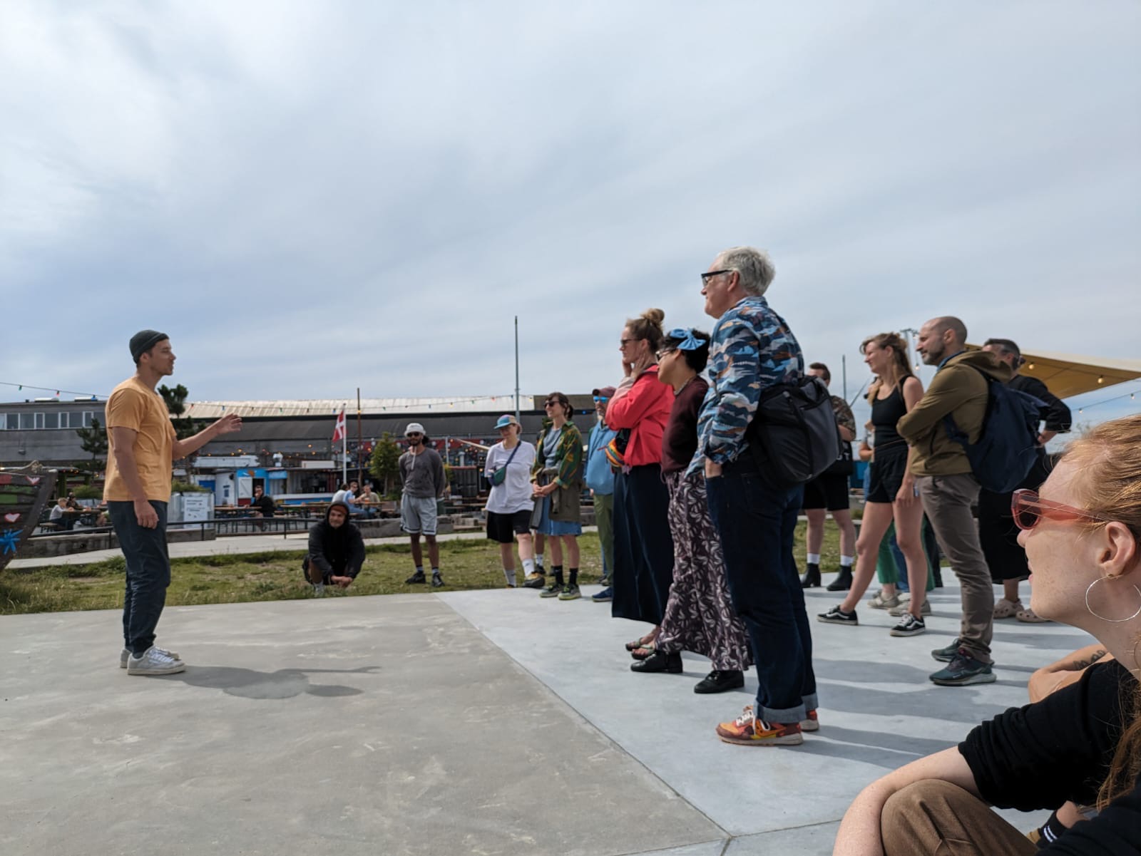 Een man in een oranje shirt spreekt een groep mensen toe op een plein met gras en beton. De groep staat in een halve cirkel en luistert geconcentreerd. Op de achtergrond zijn industriële gebouwen en terrassen te zien.