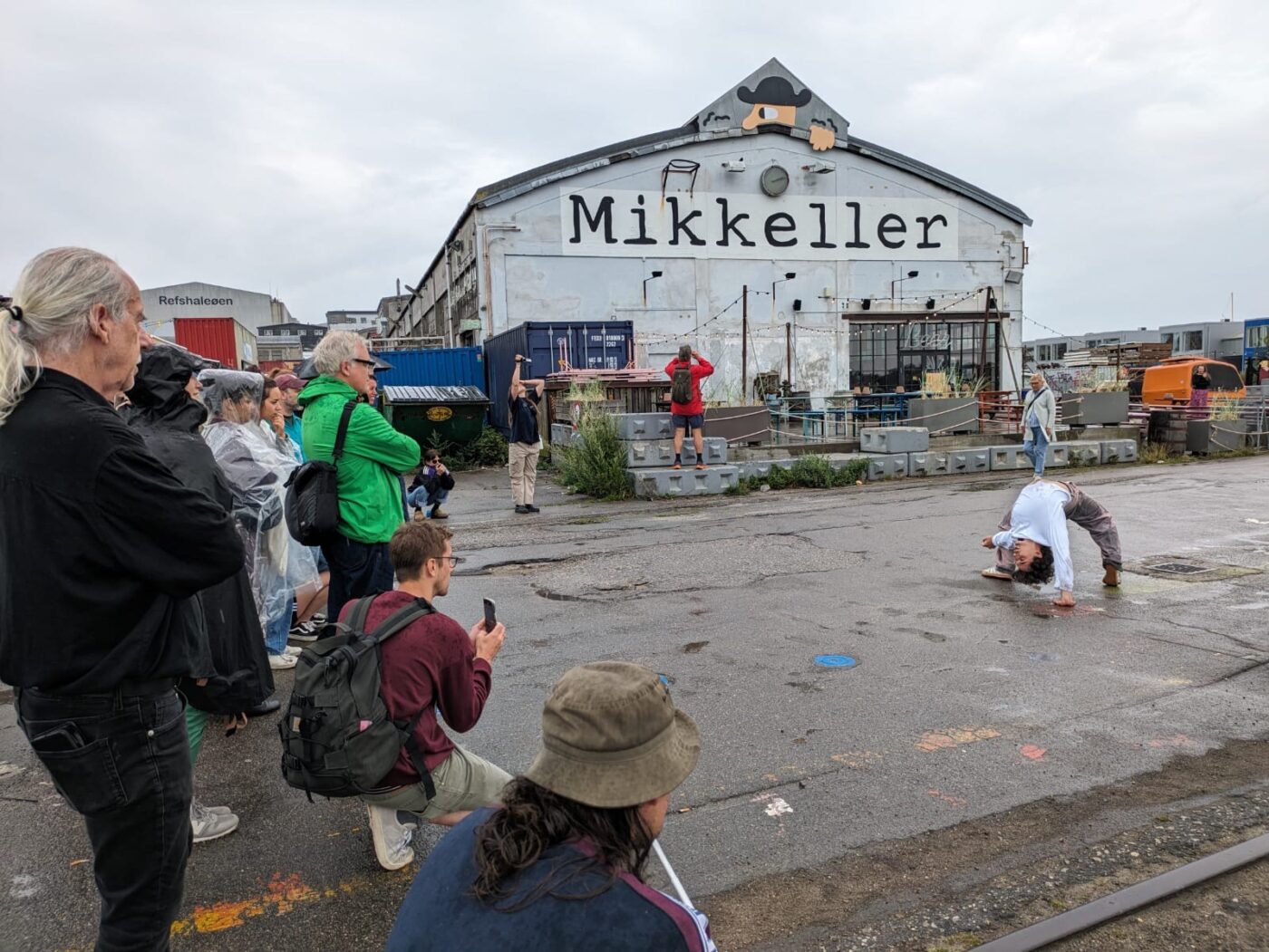 Een performer buigt achterover op een natte straat bij het Mikkeller-gebouw in een rauw, industrieel gebied. Een groep toeschouwers, sommigen in regenkleding, kijkt aandachtig toe.