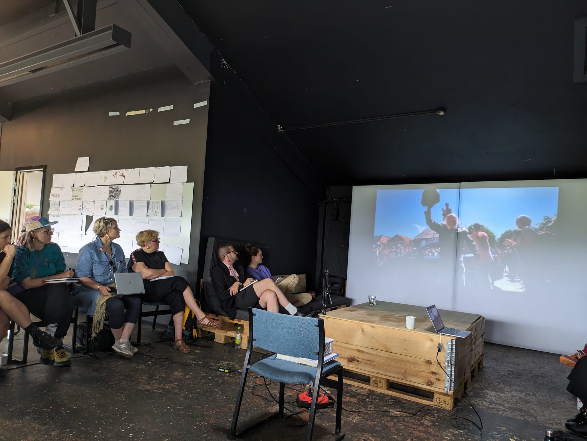 Een groep mensen zit in een donkere ruimte en kijkt aandachtig naar een presentatie die op een groot scherm wordt geprojecteerd. Op het scherm is een foto te zien van een buitenvoorstelling met muzikanten of performers. Op de muur hangen schetsen en aantekeningen.