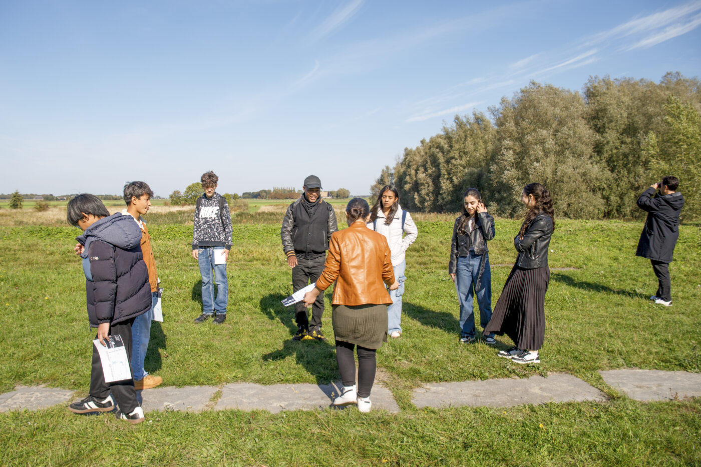Een groep jongeren staat in een kring op een open grasveld onder een blauwe hemel. Een begeleider in een oranjebruin jasje spreekt de groep toe. Enkele leerlingen kijken om zich heen of maken aantekeningen. Op de achtergrond staan bomen en is het uitgestrekte landschap zichtbaar.