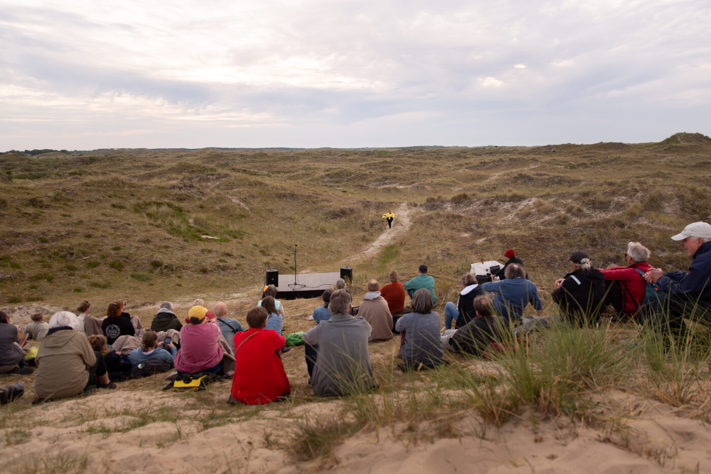 Een groep mensen zit in de duinen op zand en gras, gericht naar een klein podium met een microfoon. In de verte loopt een performer in een felgele jas over het pad richting het podium, onder een licht bewolkte hemel.
