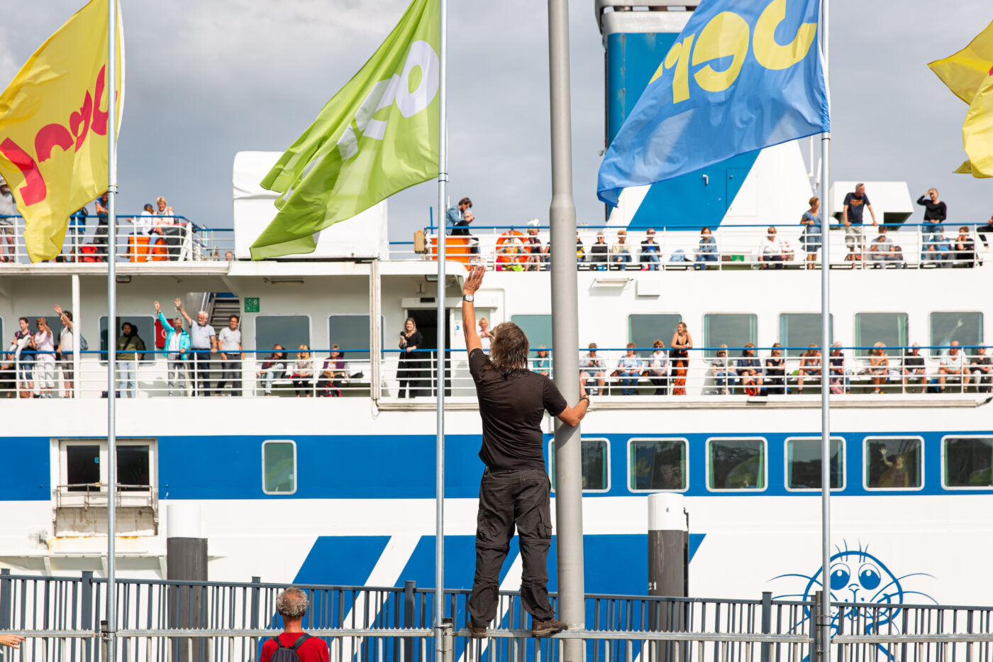 Een man zwaait vanaf de kade naar een volle veerboot waar mensen aan dek terugzwaaien. Tussen de kleurrijke vlaggen met Oerol-logo wappert de wind, terwijl de boot met blauwe en witte strepen gereedstaat voor vertrek.
