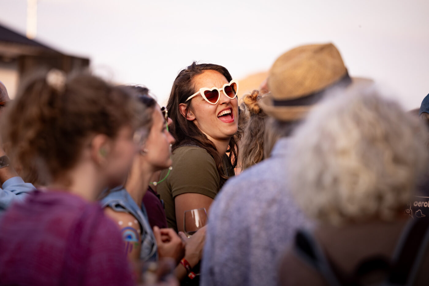 Een groep festivalbezoekers staat dicht op elkaar in de buitenlucht. Centraal valt een lachende vrouw op met lange haren en opvallende hartvormige zonnebril; ze straalt plezier uit te midden van de kleurrijke, zomerse menigte.